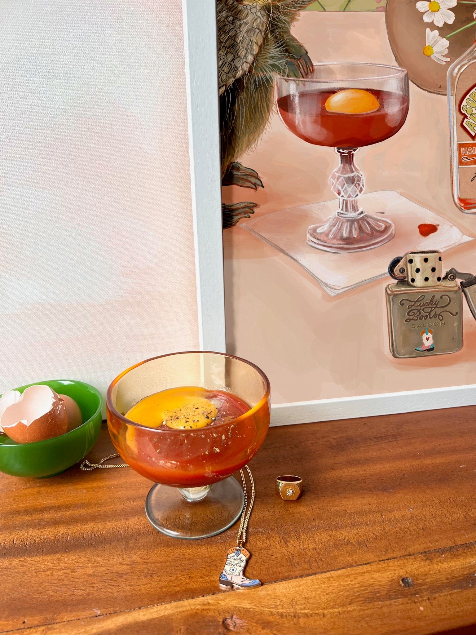 The Prairie Oyster Giclée Art Print by Hôtel Dodo depicts a cocktail with raw egg yolk, resting on wood beside a bowl of eggshells and jewelry—a stylish homage to the classic hangover cure, ideal for any gift shop display.