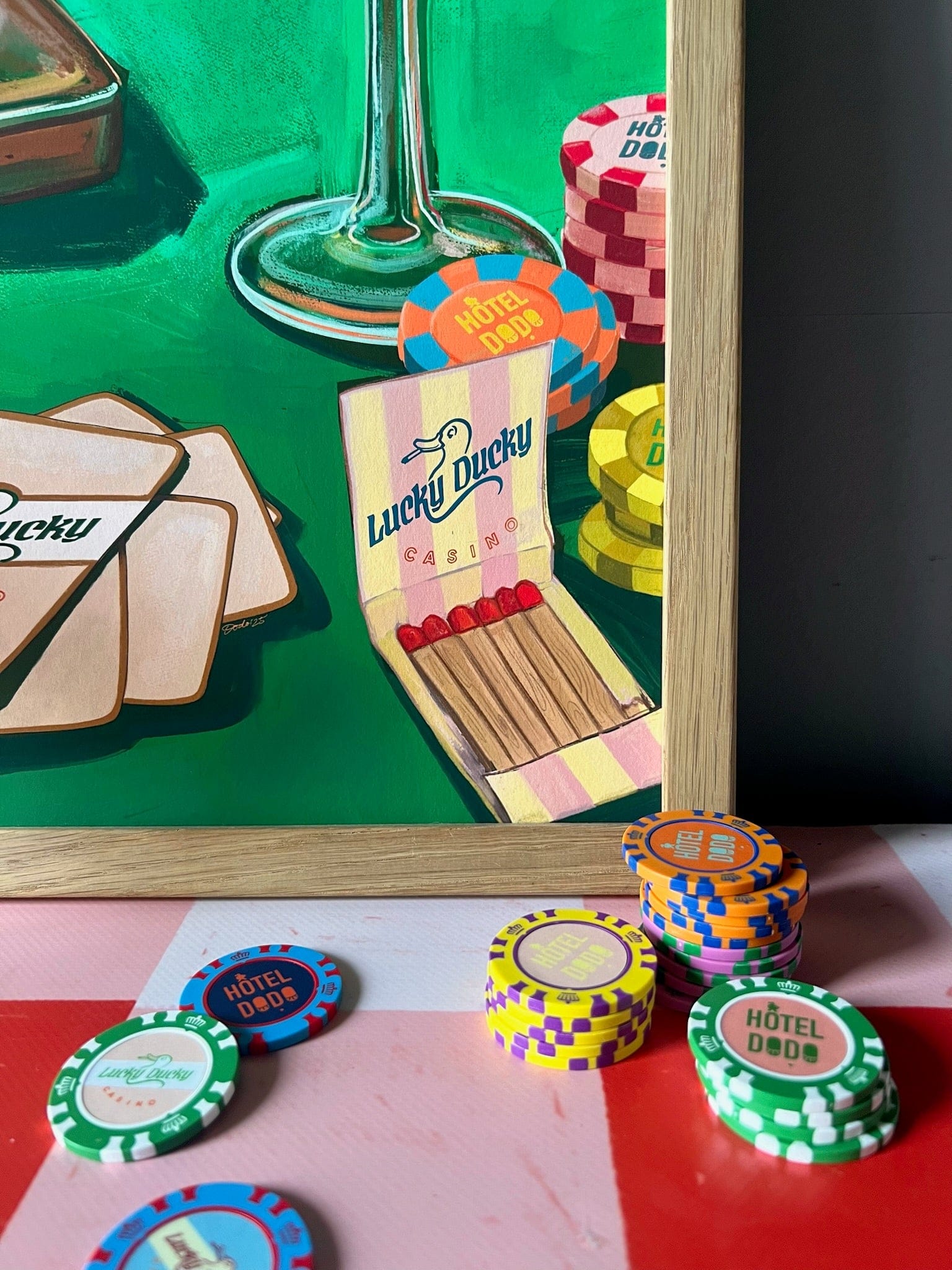 A close-up of poker chips and cards on a table, with a "Lucky Ducky Casino Giclée Art Print" by Hôtel Dodo next to a matchbook and the base of a wine glass, adding playful casino flair to the scene.