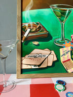 The Lucky Ducky Casino Giclée Art Print by Hôtel Dodo features playing cards, cigarettes, and martinis atop a green table, displayed next to a real martini glass and poker chips on a red-and-white checkered surface.