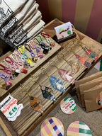 A wooden display at a market stall features Hôtel Dodo’s Bodega Cat Hot Sauce Keyring, colorful charms, enamel motel-inspired tags, business cards, and small envelopes arranged neatly on the table.