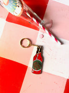 The Hôtel Dodo Aperitivo Keyring, shaped like a vintage soda bottle, sits on a red and white checkered surface beside a spilled glass with pink liquid—a quirky charm perfect for adding flair to your bag.