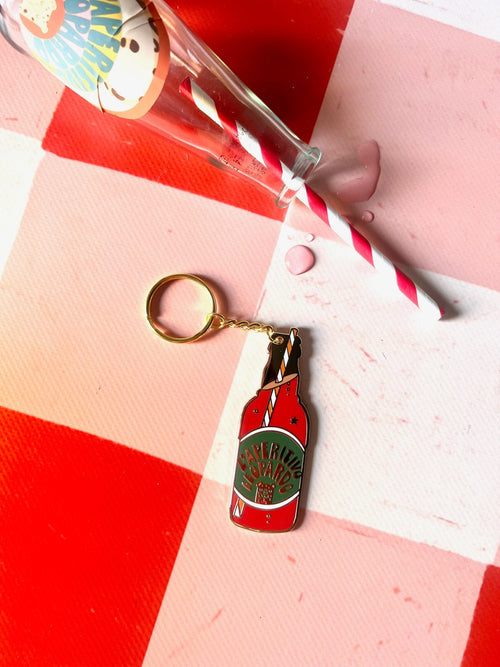 The Hôtel Dodo Aperitivo Keyring, shaped like a vintage soda bottle, sits on a red and white checkered surface beside a spilled glass with pink liquid—a quirky charm perfect for adding flair to your bag.
