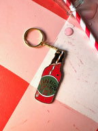 The Hôtel Dodo Aperitivo Keyring is a quirky bag charm shaped like a red vintage soda bottle with a striped straw and "PAPERITO LEOPARDO" label, shown on a pink and red patterned surface.