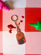 The Bodega Cat Hot Sauce Keyring by Hôtel Dodo, featuring an enamel tag, rests on a red and pink checkered surface with hot sauce drops and two squeeze bottles—one pink, one green—in the corners.