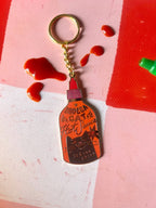 The Hôtel Dodo Bodega Cat Hot Sauce Keyring, featuring a metal keychain with a hot sauce bottle enamel tag, rests on a pink surface with red sauce drops—making it the perfect quirky bag charm.