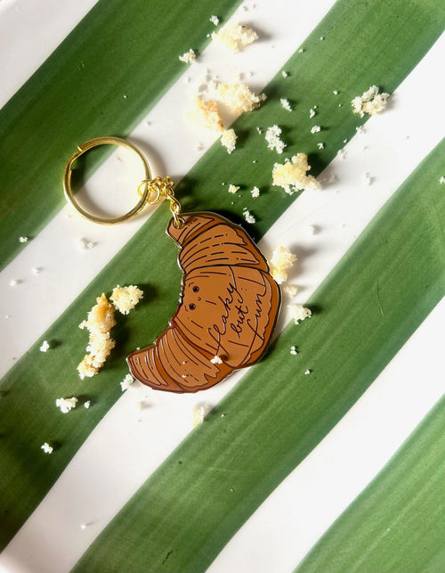 The Kwasong Keyring by Hôtel Dodo, shaped like a croissant and inscribed with "la vie est belle mon ami," sits on a green and white striped plate amid scattered crumbs.
