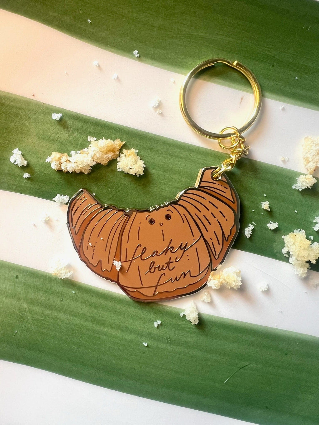 The Hôtel Dodo Kwasong Keyring, a croissant-shaped keychain with "flaky but fun" lettering and an enamel tag, rests on green-and-white stripes amid pastry crumbs—a quirky and adorable bag charm.