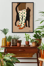 The "Encounters of the Purred Kind I" Matte Art Print by Beauty and the Beastie hangs above a wooden table with assorted potted plants, depicting a lucky black cat arching around polka-dotted boots.