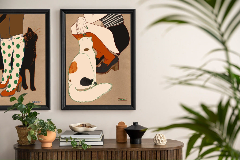 Two Encounters of the Purred Kind II Matte Art Prints by Beauty and the Beastie hang above a shelf with books, plants, and décor; a leafy plant sits in the foreground.