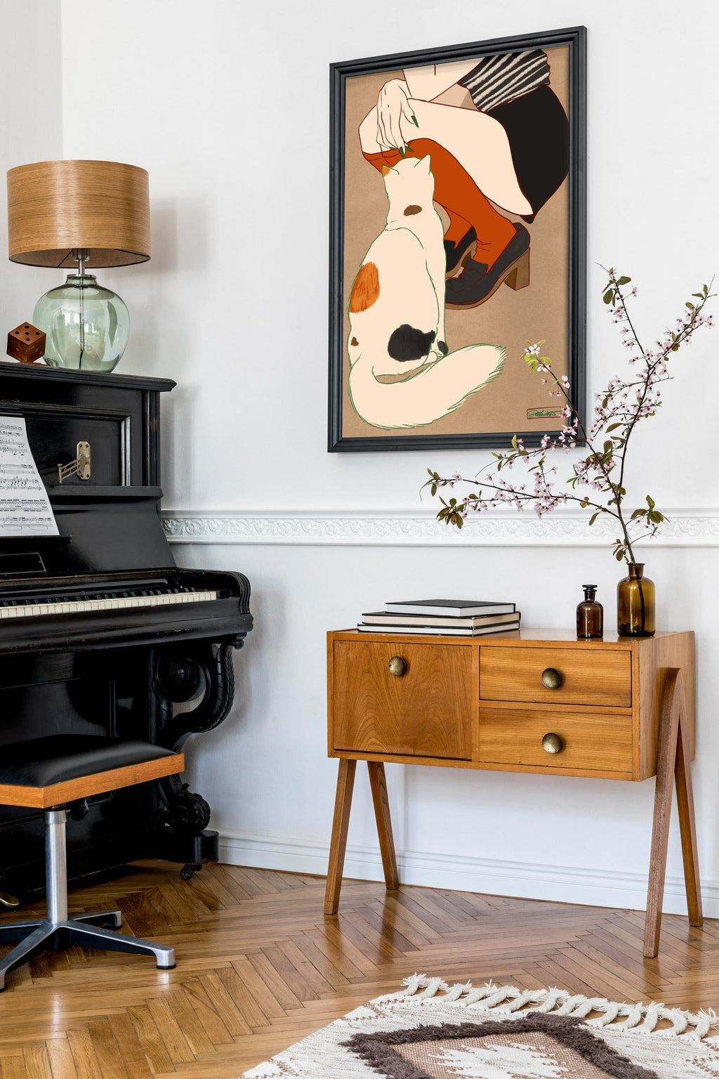 A modern living room with a black piano, wooden side table, chair, lamp, and vase with branches. On the wall hangs Encounters of the Purred Kind II Matte Art Print by Beauty and the Beastie, featuring a calico cat and person.