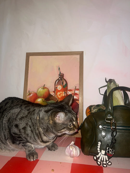 A gray tabby cat sniffs a garlic bulb on a red-and-white table, next to a green bag with a cat keychain and the Apple Core Giclée Art Print by Hôtel Dodo in the background.
