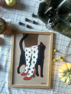 The "Encounters of the Purred Kind I Matte Art Print" by Beauty and the Beastie features a person in spotted socks and red heels with a lucky black cat, set on a checkered tablecloth with pumpkins, apples, chocolates, and a green bag.
