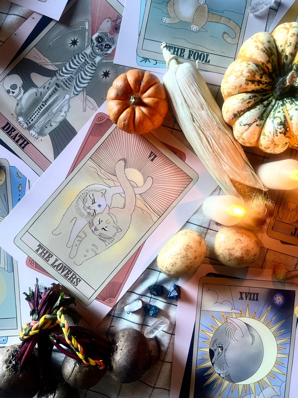 The Lovers Matte Art Print by Tarot Cats, surrounded by vegetables, small pumpkins, and a bundle of herbs on a table lit with warm string lights.