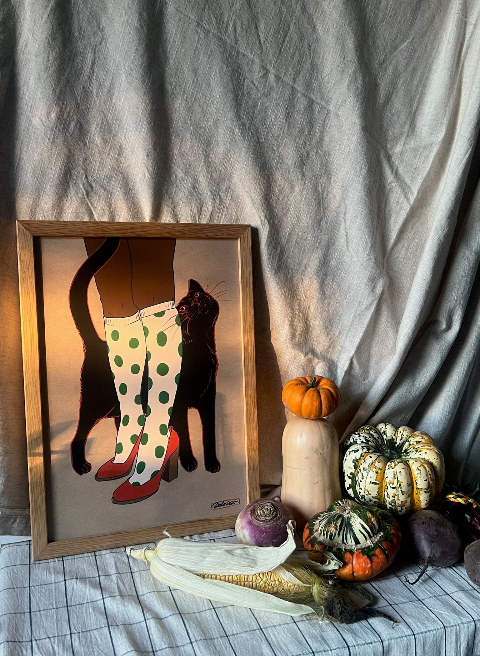 The Encounters of the Purred Kind I Matte Art Print by Beauty and the Beastie features a lucky black cat and polka dot boots, shown framed on a draped table with assorted squash, gourds, and root vegetables.
