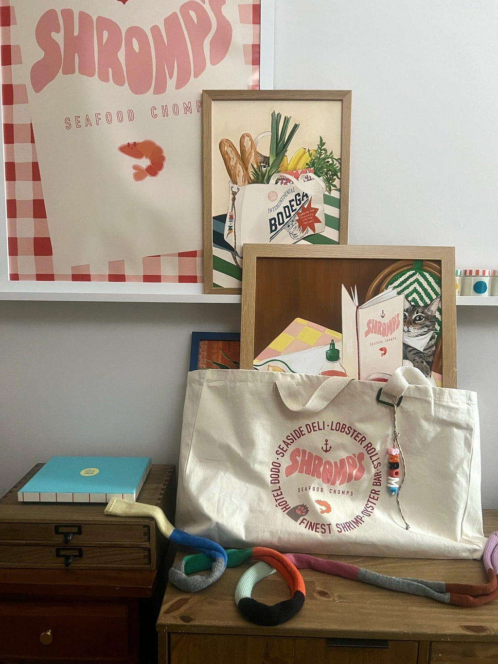 The Bodega/Shromp’s XXL Snack Shopper by Bags by diedododa, an eco-friendly cotton canvas tote featuring a "Shromp's Seafood Chowder" logo, sits on a dresser with framed art, a blue notebook, and a colorful knit chain—stylish and practical!.