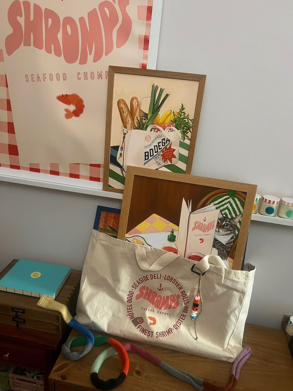 The Bags by diedododa Bodega/Shromp’s XXL Snack Shopper sits on a shelf by colorful items, a blue box, framed food-themed art, and a poster, its "Shromps" logo clearly visible on the cotton canvas.
