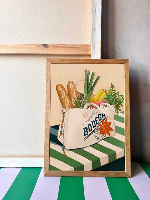 The Bodega Bounty Giclée Art Print by Hôtel Dodo features a framed painting of a grocery bag with baguettes, produce, and herbs on green-and-white stripes, capturing the simple pleasures of everyday life.