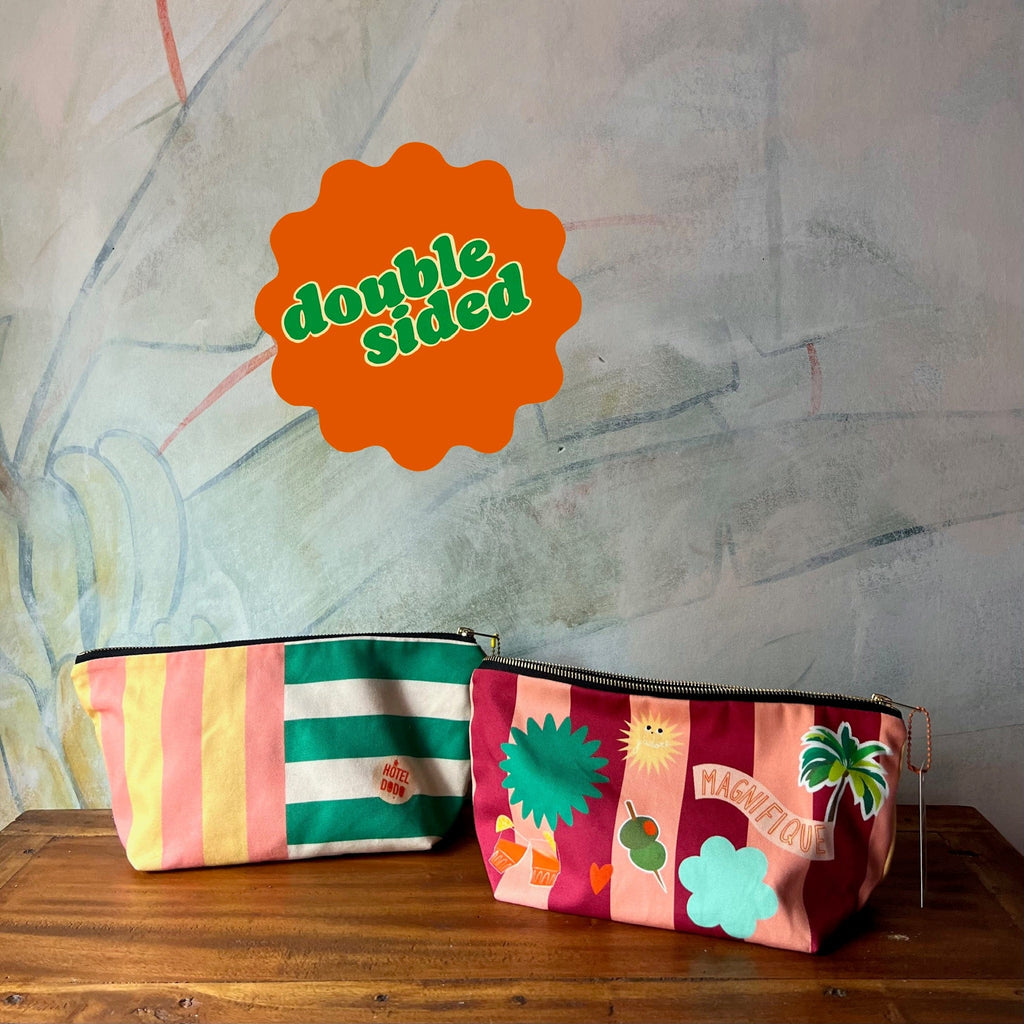 Two zippered Patch Pick ‘n’ Mix Pouches by Hôtel Dodo, featuring colorful stripes and playful designs, rest on a wooden surface with a double-sided orange and green sign in the background. Handmade in the UK, they're the perfect versatile organiser pouch.
