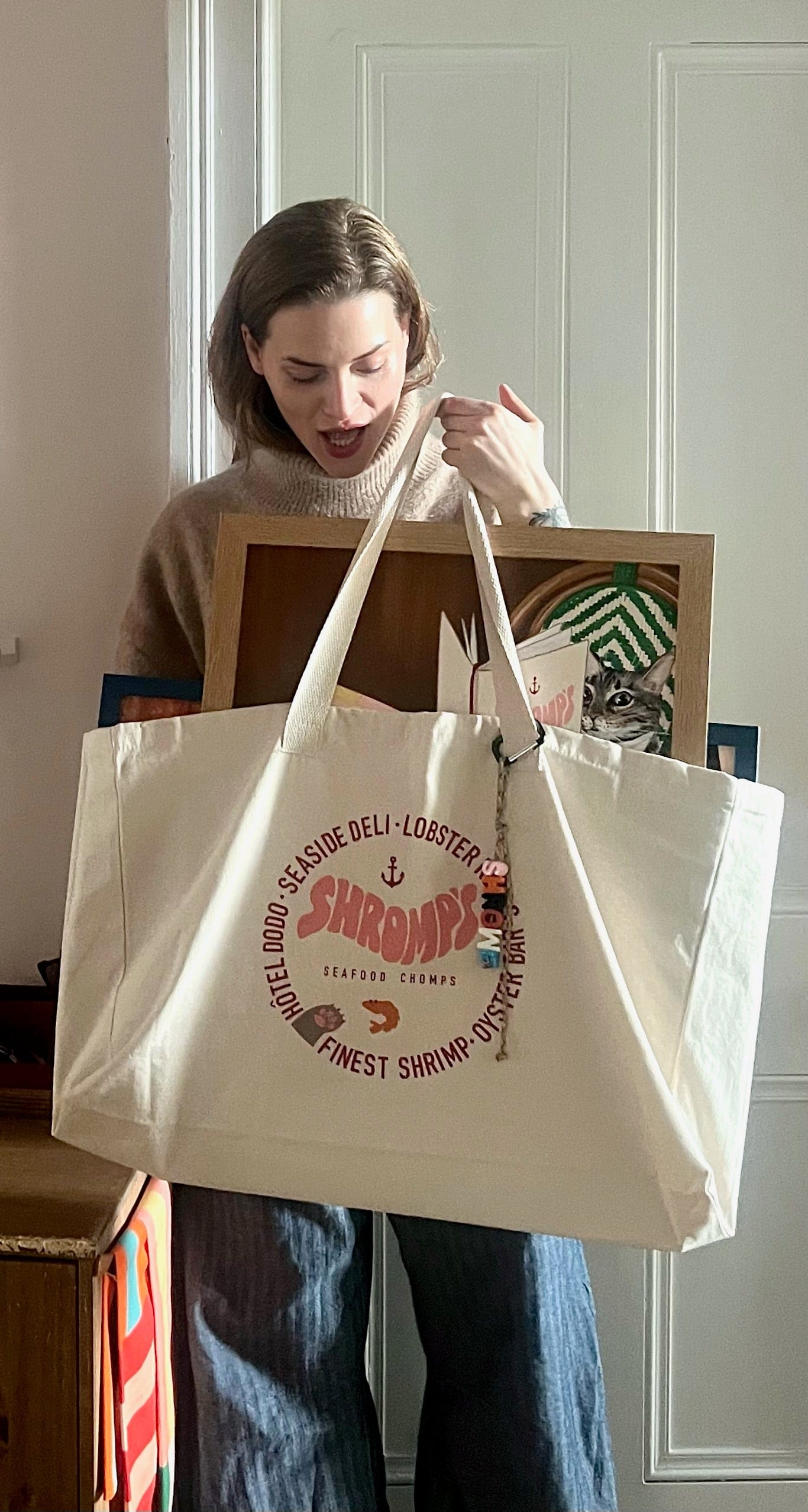 A woman indoors holds the Bodega/Shromp’s XXL Snack Shopper by Bags by diedododa, an eco-friendly cotton canvas bag, while carrying two framed items under her arm.