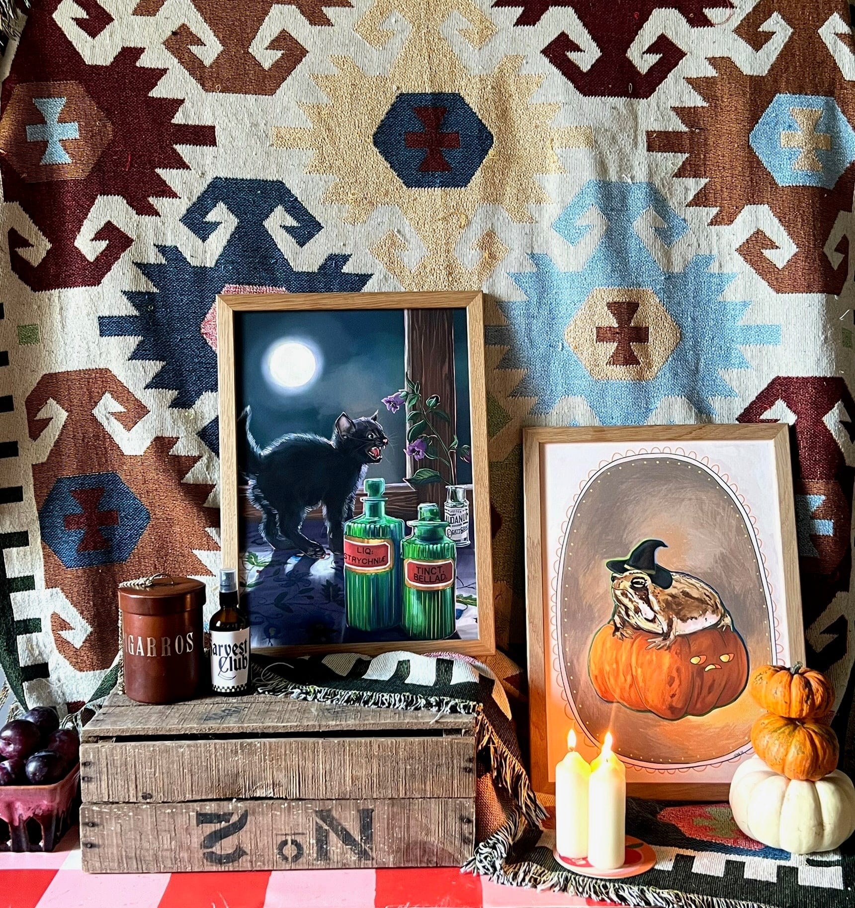 A still life featuring Kitsch Kanaveral’s The Potionist's Apprentice Giclée Art Print, surrounded by candles, pumpkins, a vintage potion bottle, plums, and crates against a textile backdrop celebrating the season.