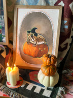 The Grumpy Spiced Pumpkin Giclée Art Print by Kitsch Kanaveral features a witchy rain frog atop a pumpkin—ideal décor for cozy Halloween vibes.