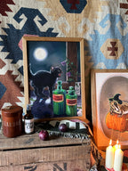 The Potionist's Apprentice Giclée Art Print by Kitsch Kanaveral features a black cat with green bottles on a trunk amid candles, onions, and artwork, conjuring an All Hallows’ Eve mood before a rich tapestry backdrop.