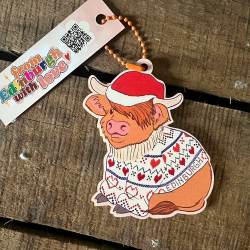The Christmas by diedododa Christmas Cow Hanging Decoration features a cartoon Highland cow in a festive sweater and Santa hat labeled "Edinburgh," displayed on a rustic wooden surface.