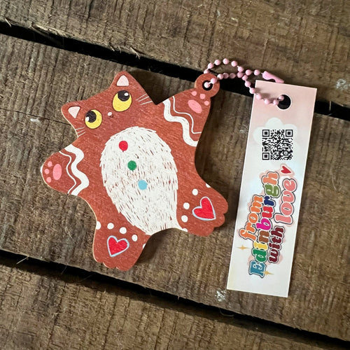 The GingerBread Cat Hanging Decoration by Christmas by diedododa is a wooden cat-shaped keychain with colorful painted details, a "from Edinburgh with love" tag, and a colored short metal chain, displayed on a wooden surface.