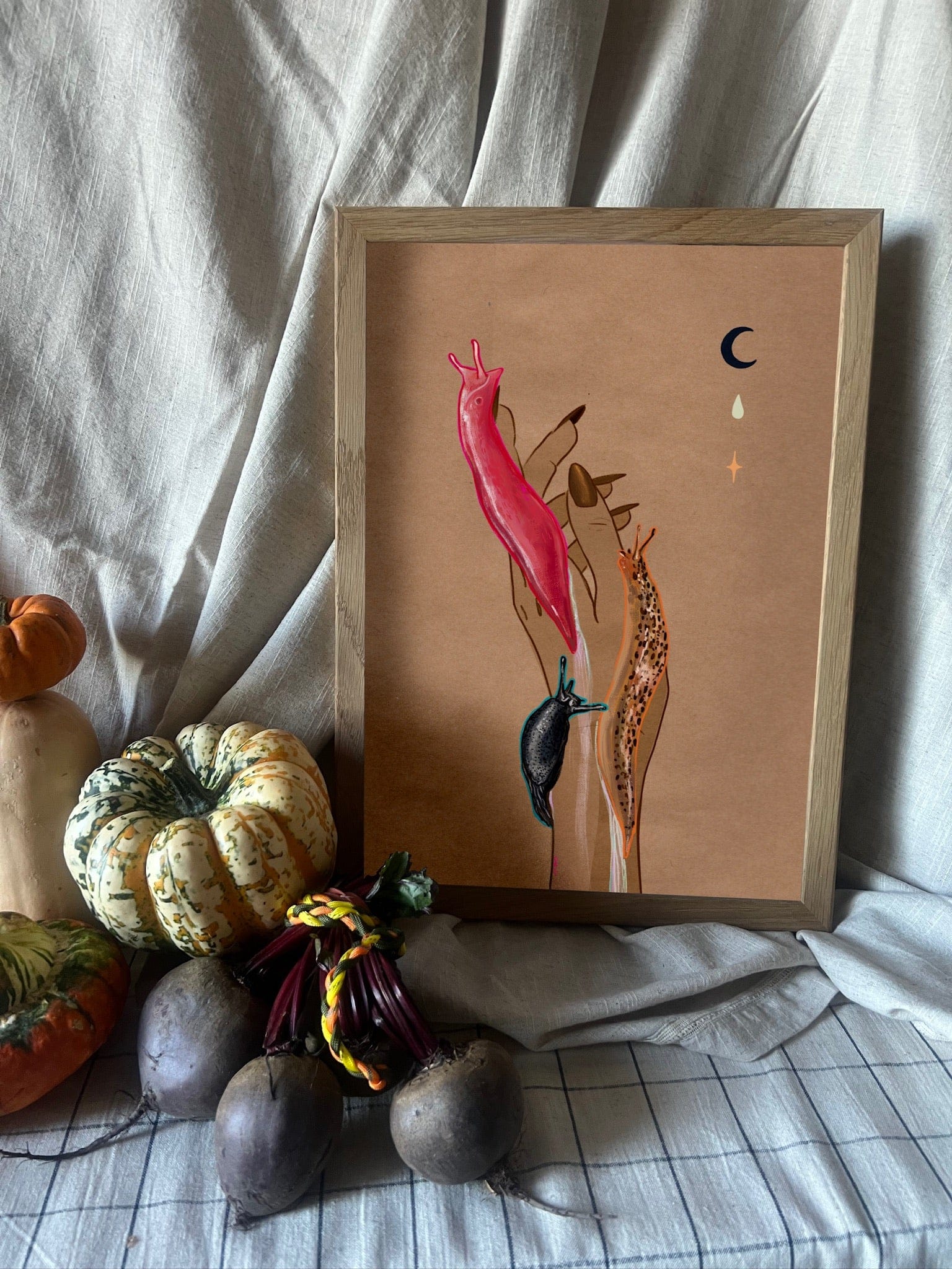 The Midnight Slugs Matte Art Print by Beauty and the Beastie features hands with vibrant leopard and Australian pink slugs on the fingers, surrounded by gourds and beets against a neutral draped fabric background.