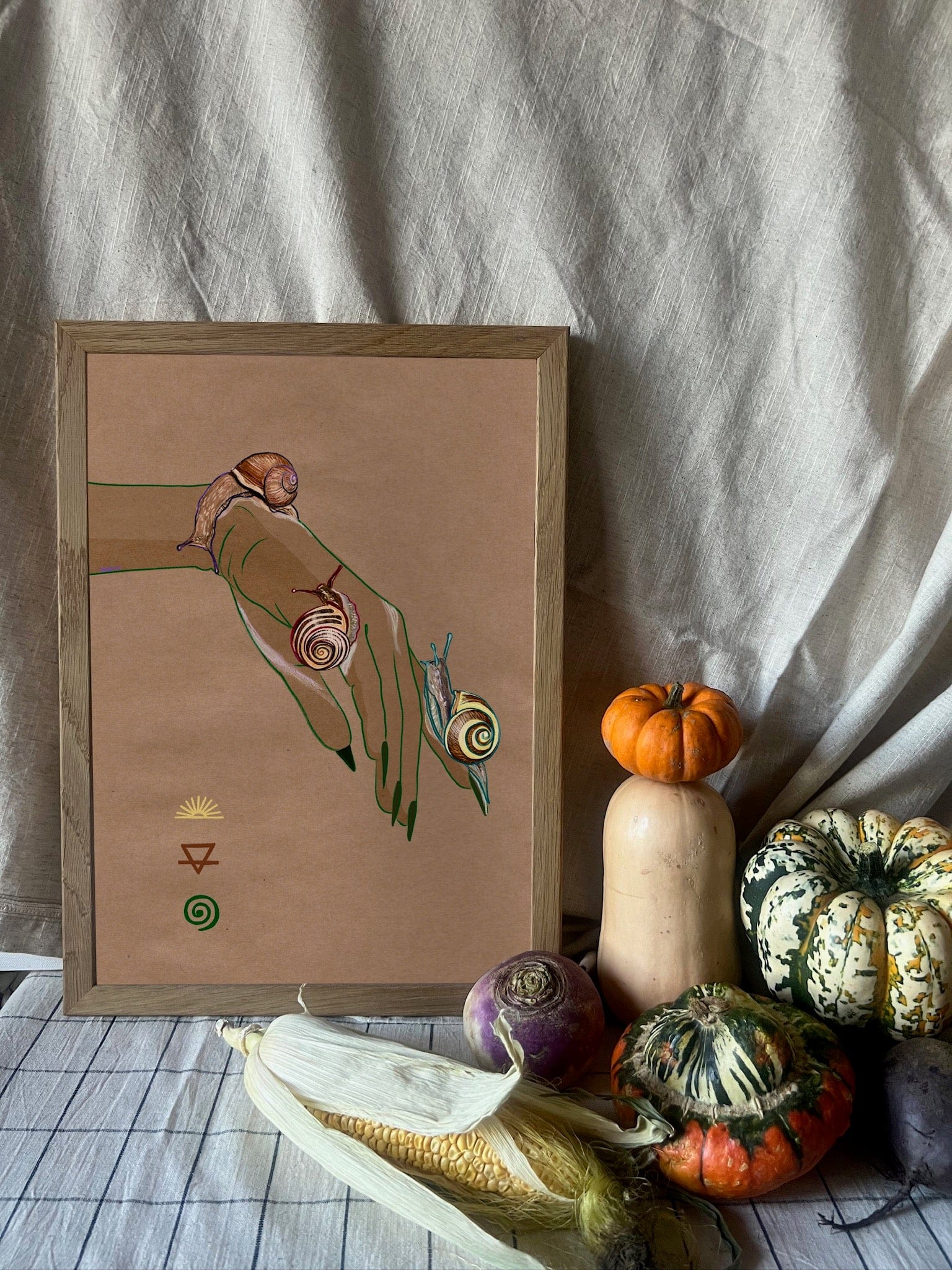 The Spring Snails Matte Art Print by Beauty and the Beastie features a framed hand with snails, set on a table with gourds, corn, and turnip, all against a neutral fabric backdrop.