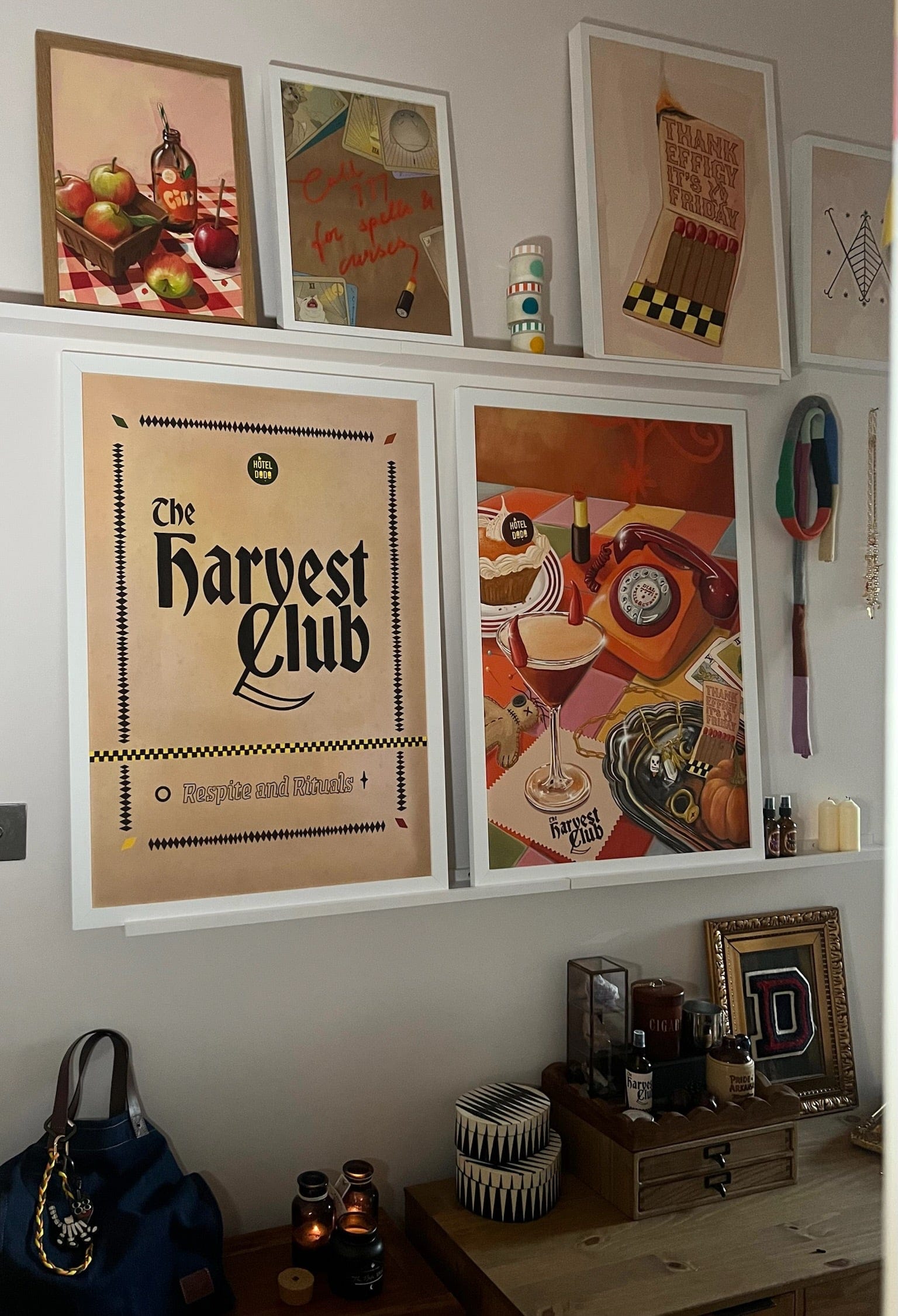 A cozy room corner features The Harvest Club Giclée Art Print by Hôtel Dodo on white shelves, surrounded by decor items, candles, and bags on a wooden surface—creating a subtle spooky-season vibe.