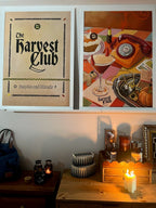 Two framed "The Harvest Club Giclée Art Print" by Hôtel Dodo hang above a wooden dresser, their paranormal vibe pairing perfectly with candles, books, and bottles—ideal decor accents for the spooky season.