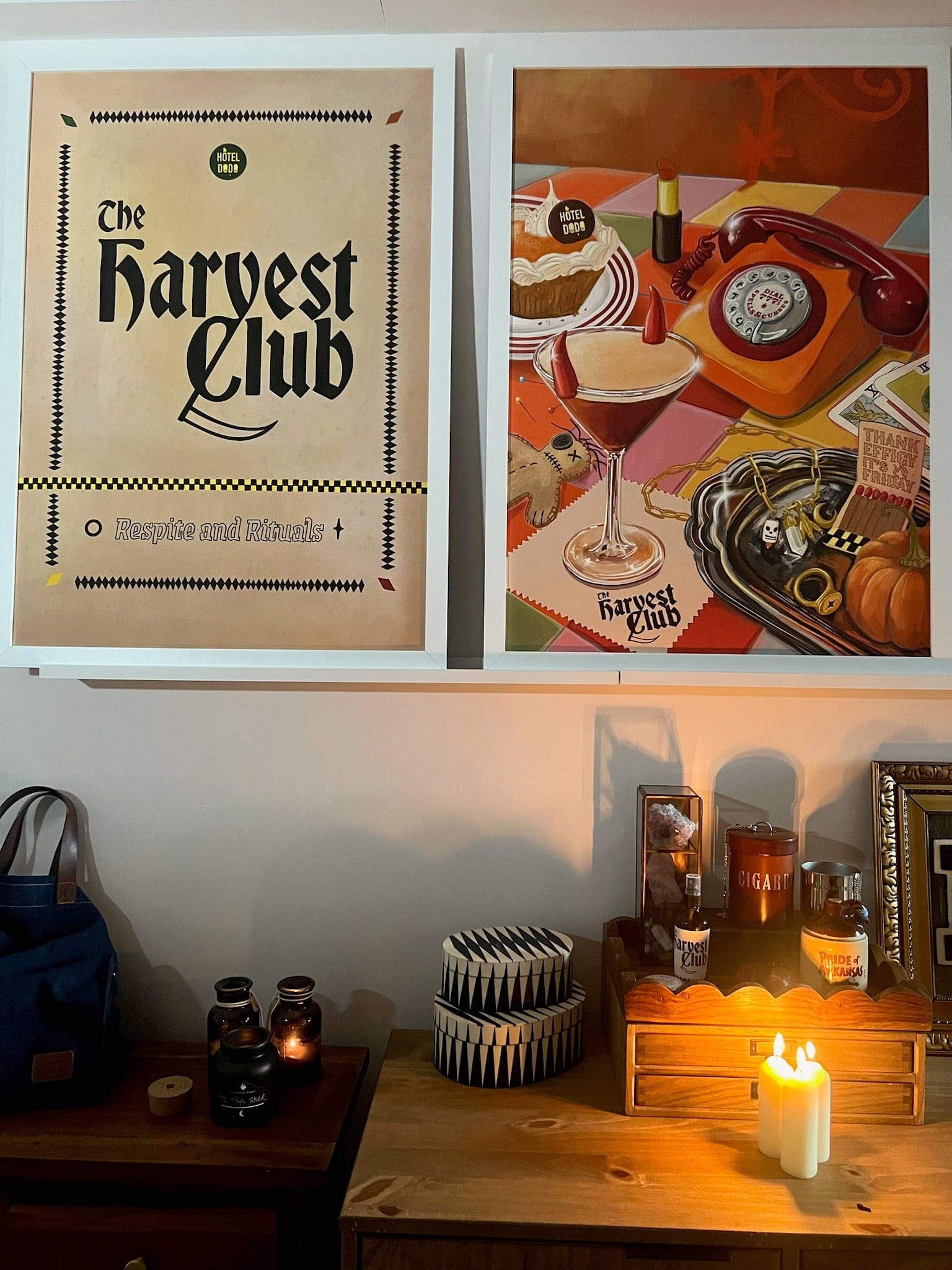 Two framed "The Harvest Club Giclée Art Print" by Hôtel Dodo hang above a wooden dresser, their paranormal vibe pairing perfectly with candles, books, and bottles—ideal decor accents for the spooky season.