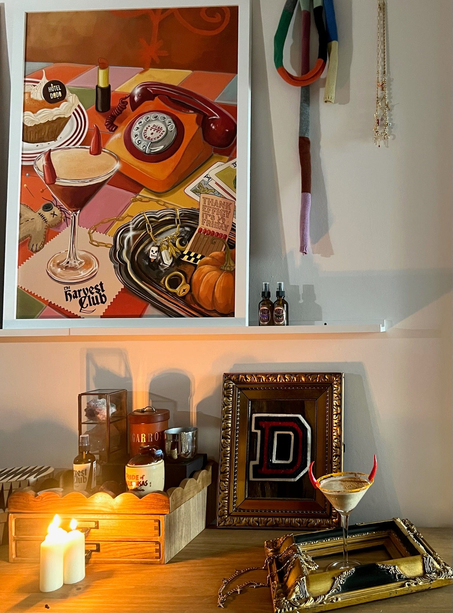 A cocktail in a gold-rimmed glass sits amid candles, vintage decor, and the Harvest Club Giclée Art Print by Hôtel Dodo, whose vivid colors enhance the space’s subtle paranormal aura.
