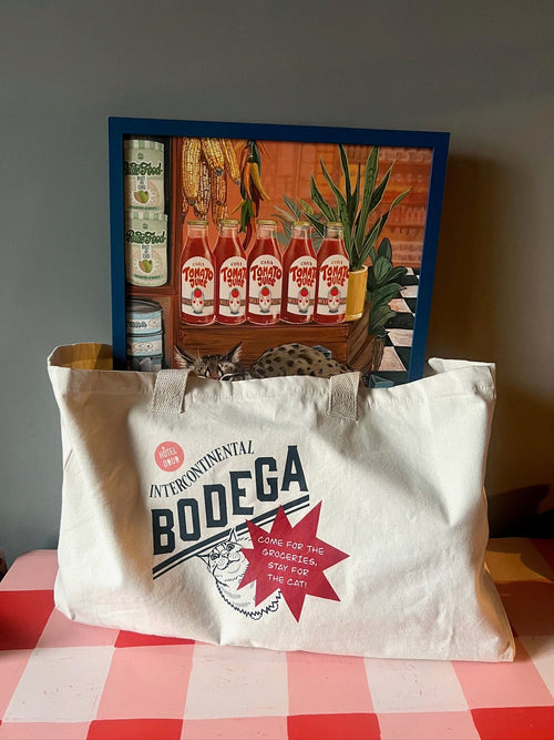 The Bodega/Shromp’s XXL Snack Shopper by Bags by diedododa, an eco-friendly white cotton canvas bag, rests on a red-and-white table before a framed painting of tomato cans, plants, and a hidden cat.