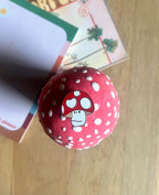 The Luckshroom Pin by Pins by diedododa—a red, white-spotted pin with a cartoon mushroom face inspired by Amanita Muscaria—rests on a wooden surface near some cards, evoking feelings of good fortune and protection.