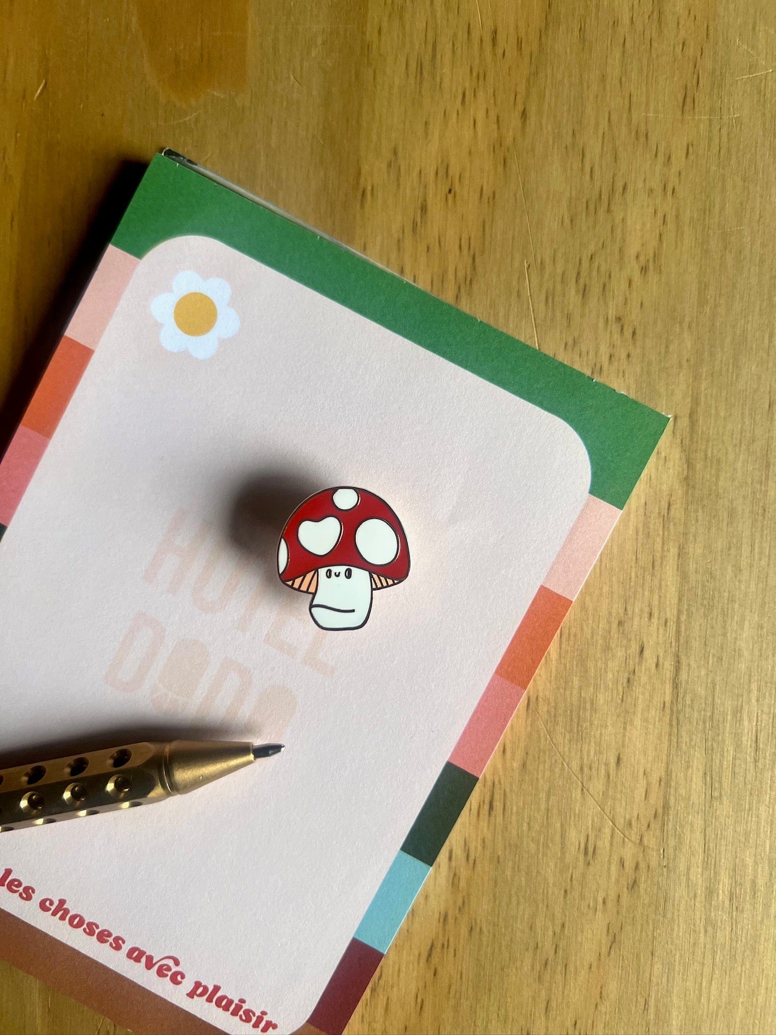 The Luckshroom Pin by Pins by diedododa, a red and white Amanita Muscaria mushroom pin symbolizing good fortune, rests on a notepad with a colorful border and flower illustration beside a gold pen on a wooden surface.