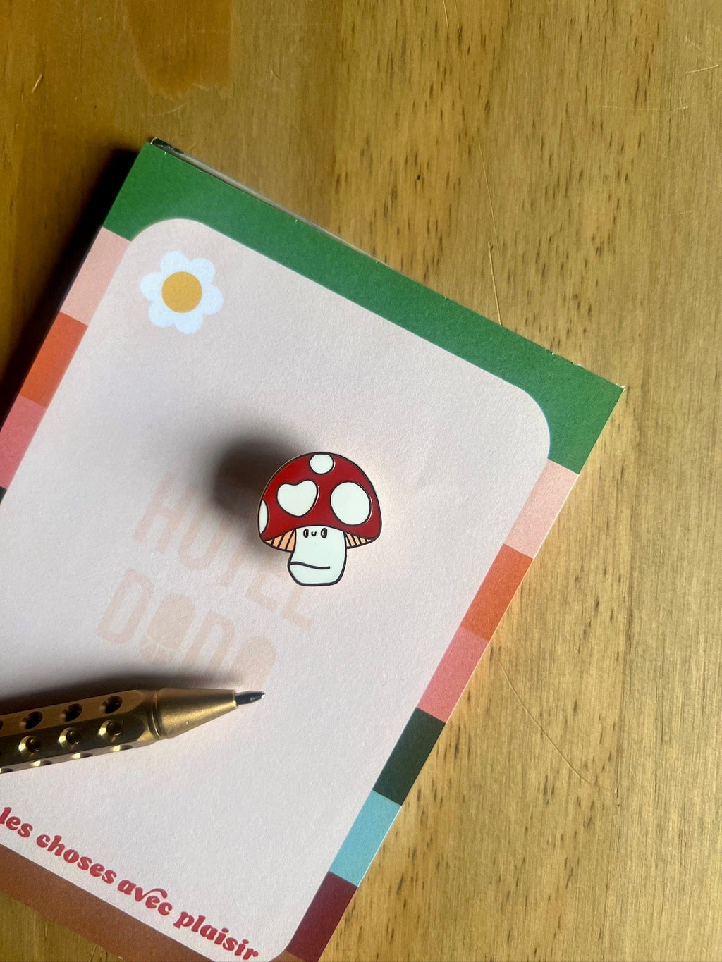 The Luckshroom Pin by Pins by diedododa, a red and white Amanita Muscaria mushroom pin symbolizing good fortune, rests on a notepad with a colorful border and flower illustration beside a gold pen on a wooden surface.