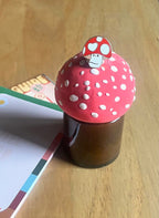 The Luckshroom Pin by Pins by diedododa features a brown glass jar with a red, Amanita Muscaria-inspired mushroom cap lid and a mushroom pin—designed to bring protection and good fortune.