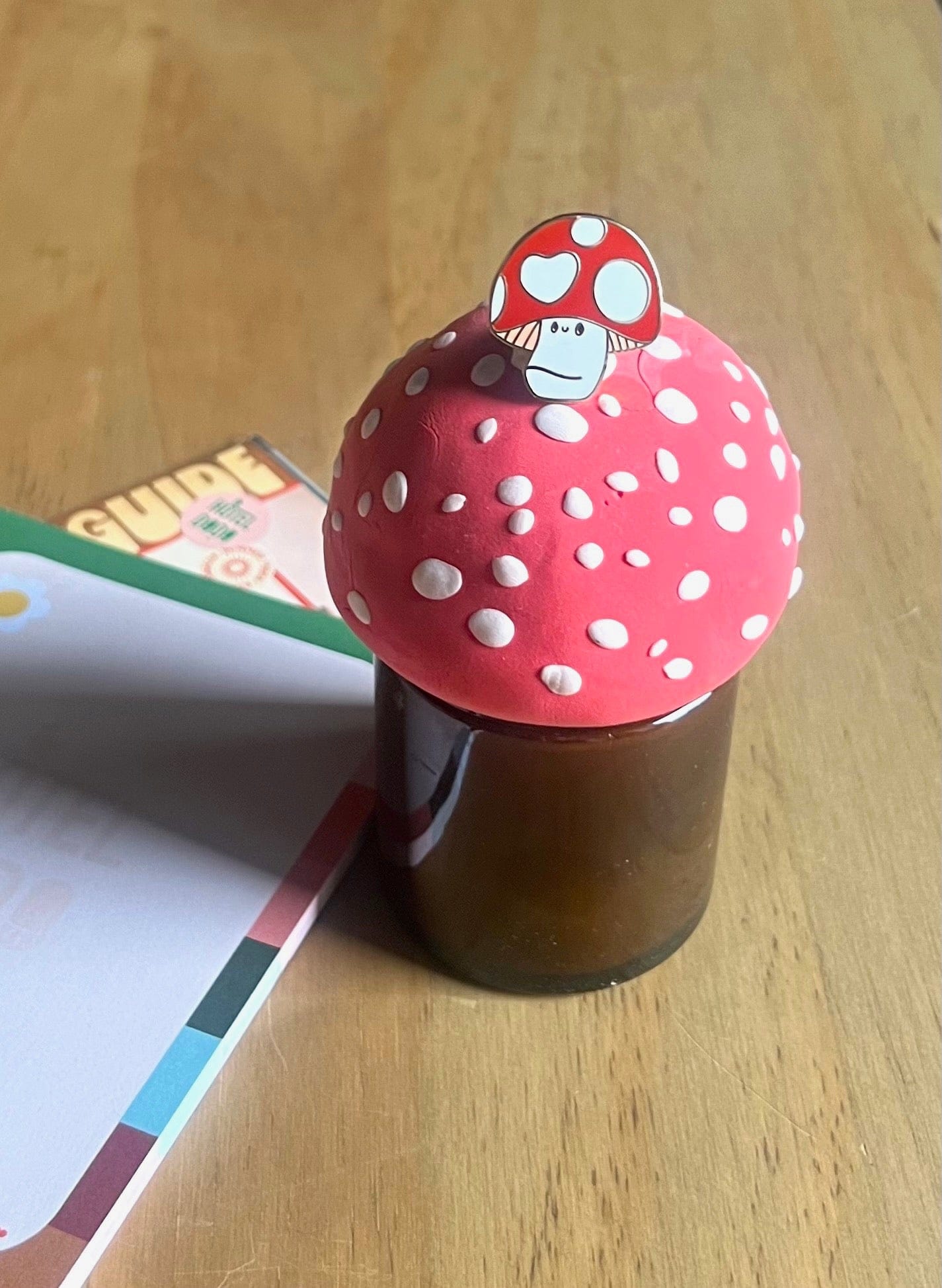 The Luckshroom Pin by Pins by diedododa features a brown glass jar with a red, Amanita Muscaria-inspired mushroom cap lid and a mushroom pin—designed to bring protection and good fortune.