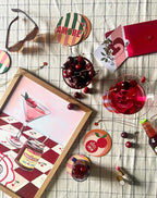A cherry-themed table featuring bowls of cherry treats, a cherry drink, coasters, sunglasses, a red wallet on a checkered cloth, and the Hôtel Dodo Cherry Bomb Giclée Art Print—like a mind palace at Hôtel Dodo.