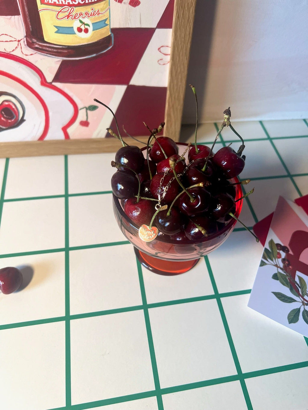 A small red glass of fresh cherries sits on a white and green grid table at Hôtel Dodo, beside a postcard and the Cherry Bomb Giclée Art Print.
