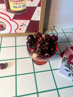 A small red glass of fresh cherries sits on a white and green grid table at Hôtel Dodo, beside a postcard and the Cherry Bomb Giclée Art Print.