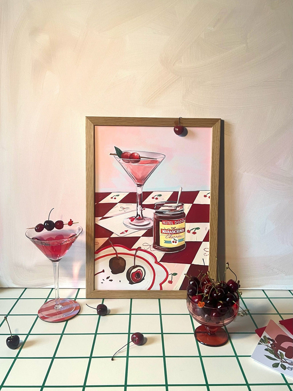 The Cherry Bomb Giclée Art Print by Hôtel Dodo features a cherry cocktail, maraschino can, and cherries on a checkered surface, evoking sweet delights beside a real cocktail glass and bowl—like treasures in a mind palace.
