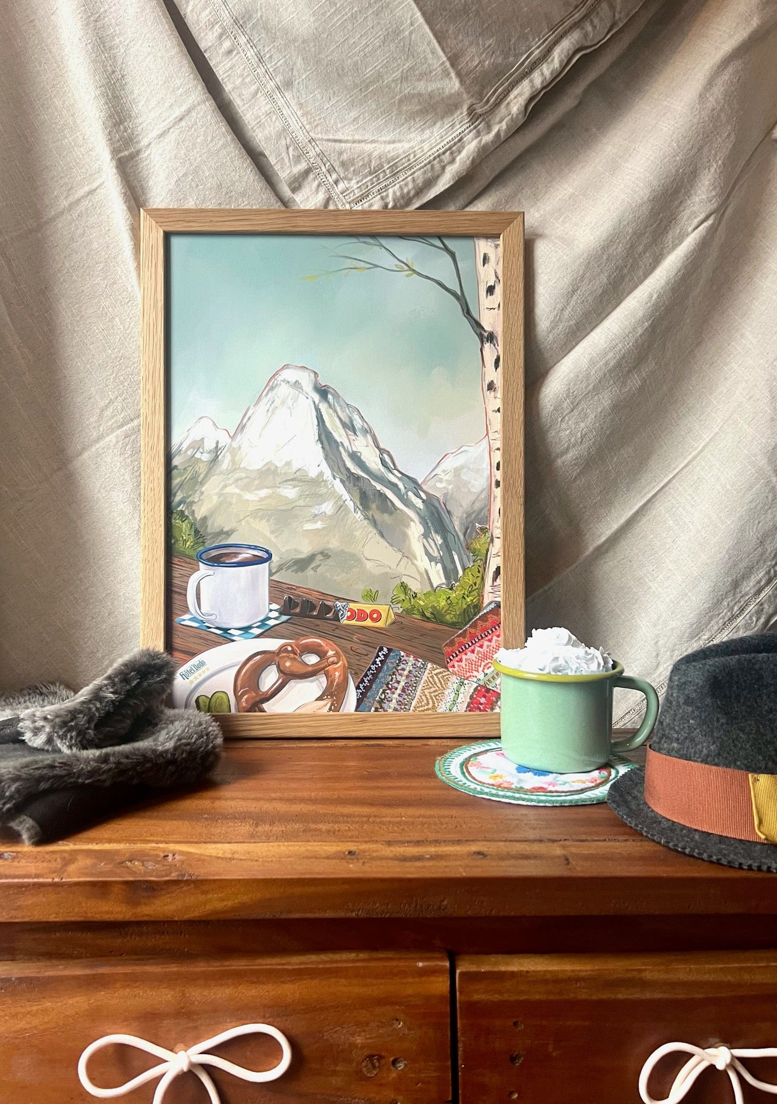 Alpine Air Giclée Art Prints by Hôtel Dodo displayed on a wooden table with a green mug, hat, faux fur, and neutral fabric—bringing the cozy charm of an alpine lodge into your space.