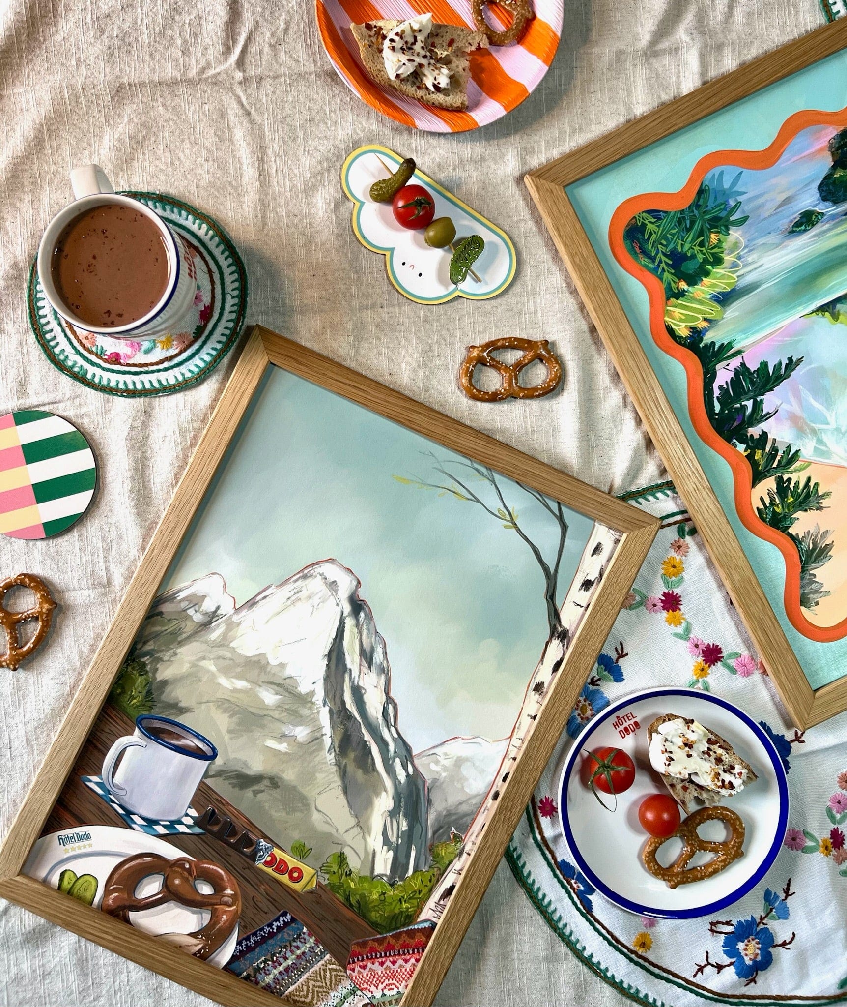 A table adorned with Kitsch Kanaveral’s Gentle Glen Giclée Art Print, plates of sweets, pretzels, tomatoes, a cup of hot chocolate, and embroidered linens atop a beige tablecloth evokes nostalgic dreamscapes.