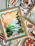 The Gentle Glen Giclée Art Print by Kitsch Kanaveral features a technicolour landscape with a wavy orange border, framed above patterned snack plates—pretzels, pickles, tomatoes, cheese—on a decorated tablecloth.