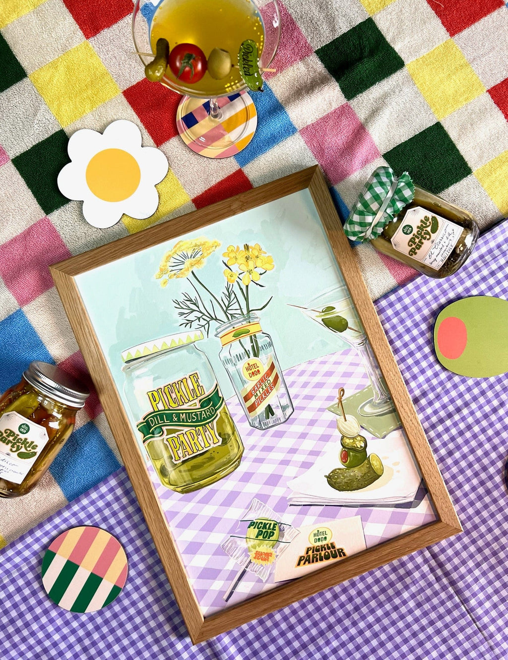 Pickle Party Giclée Art Print by Hôtel Dodo features pickle jars, flowers, drinks, and playful accents on a colorful checkered picnic blanket—spot the whimsical pickle lollipops in this charming scene!.