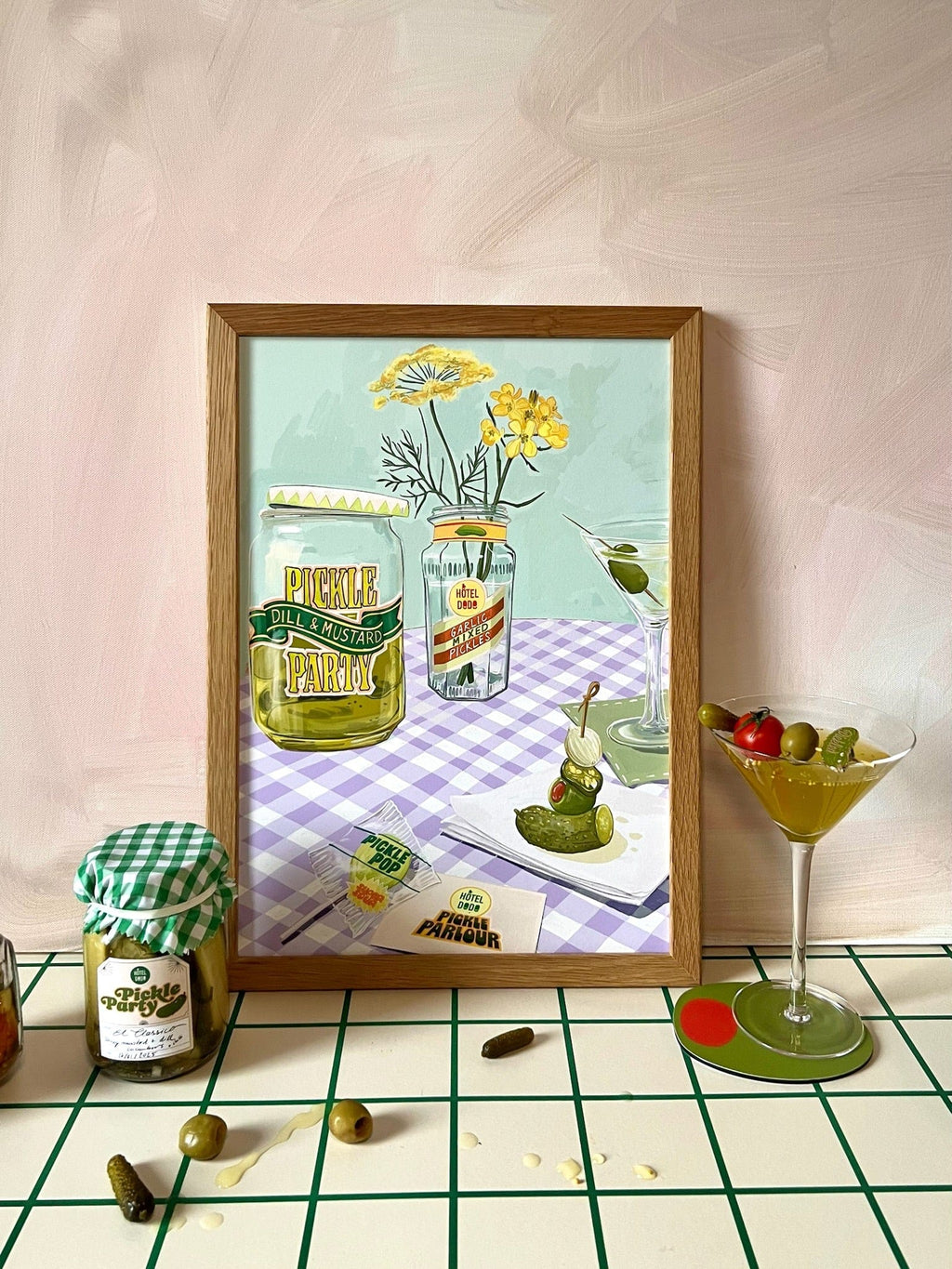 The Pickle Party Giclée Art Print by Hôtel Dodo is displayed on a table at Hôtel Dodo’s Pickle Parlour, with pickles, olives, pickle lollipops, and a martini glass arranged beside it.