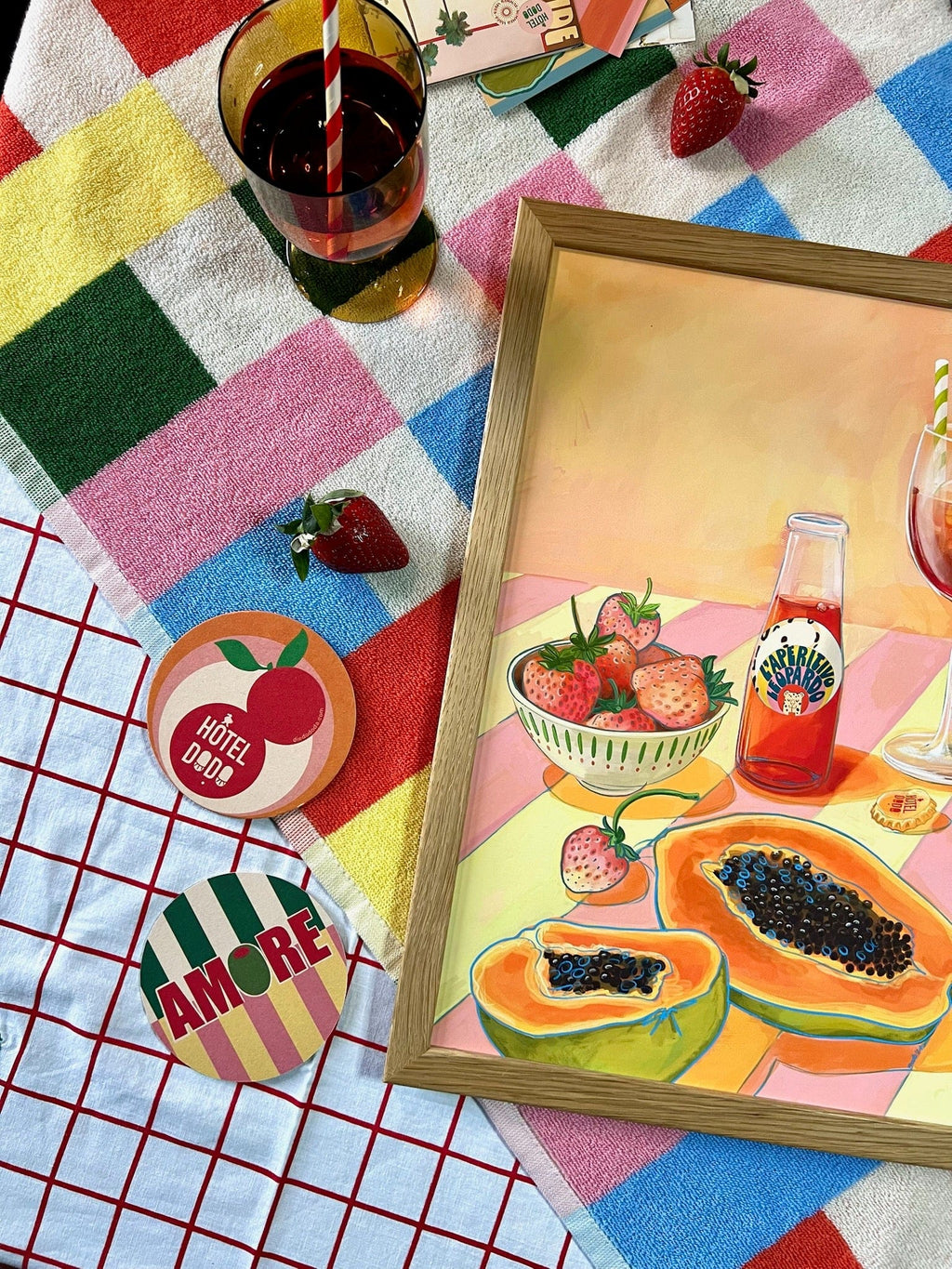 A glass of soda with a striped straw, fresh strawberries, colorful coasters, and Hôtel Dodo's L'Aperitivo Leopardo Giclée Art Print are arranged on patterned towels—capturing the playful Indian summer picnic spirit.
